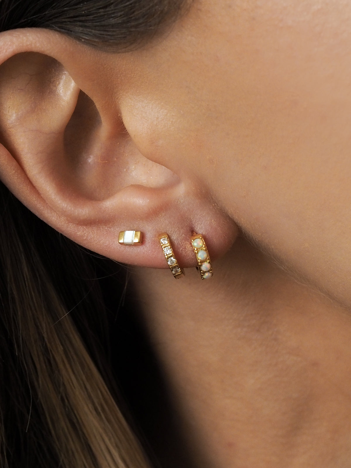 A model wearing multiple gold earrings layered together, featuring opal huggies, cubic zirconia huggies and mother of pearl studs.