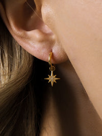 Close-up of a woman's ear wearing a gold-plated hoop earring with an eight-pointed star pendant set with small cubic zirconia stones.