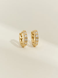 A pair of 18ct gold-plated huggie earrings with channel-set cubic zirconia stones, photographed on a soft cream background.