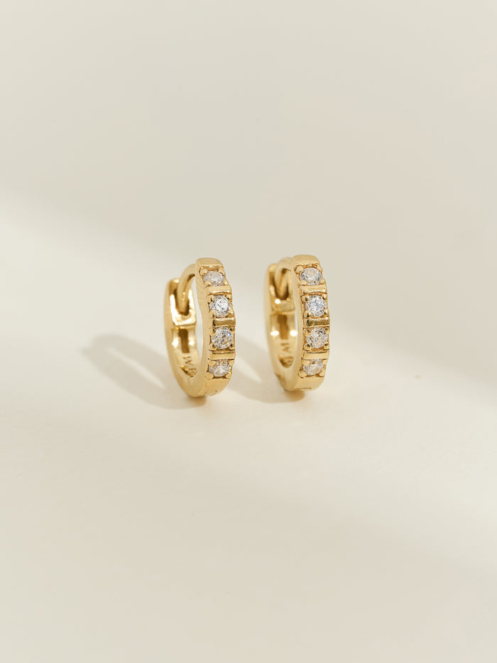 A pair of 18ct gold-plated huggie earrings with channel-set cubic zirconia stones, photographed on a soft cream background.