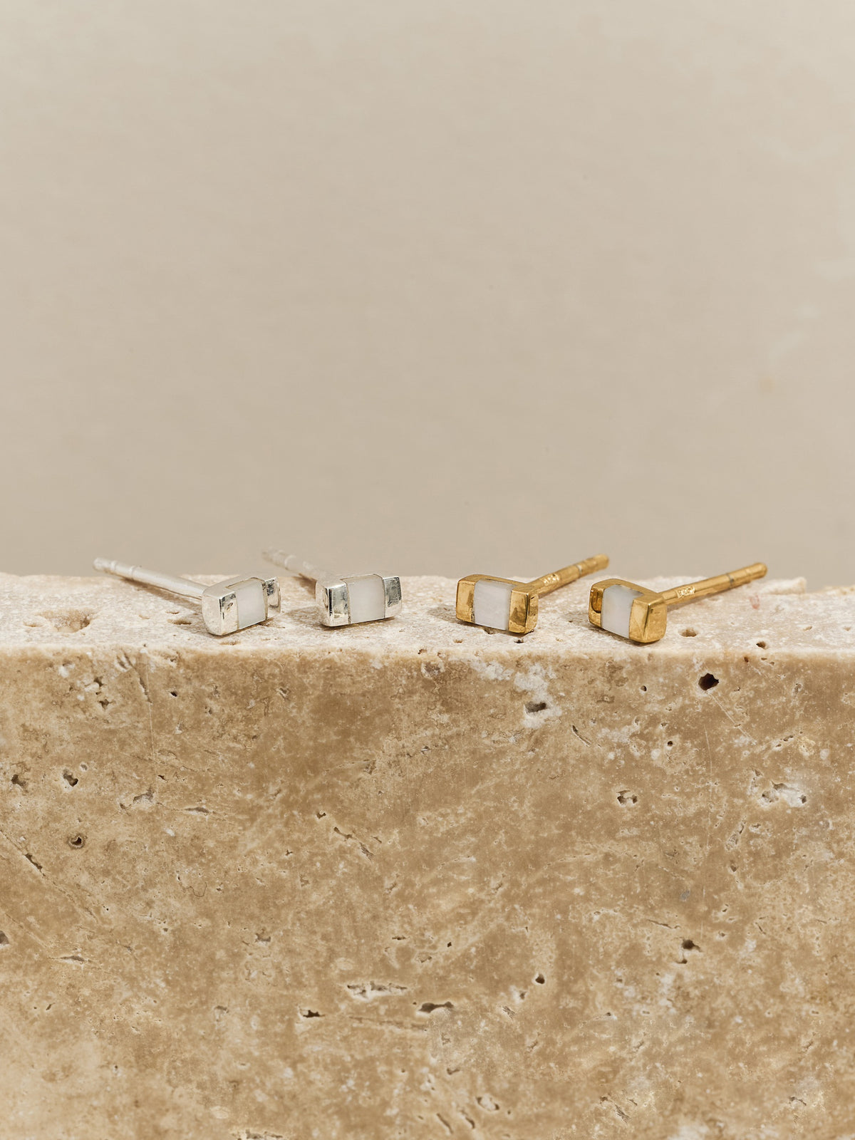 Two pairs of rectangular stud earrings with mother of pearl inlays—one in silver, one in gold—displayed on a textured stone surface.