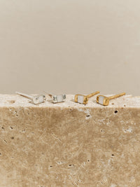 Two pairs of rectangular stud earrings with mother of pearl inlays—one in silver, one in gold—displayed on a textured stone surface.