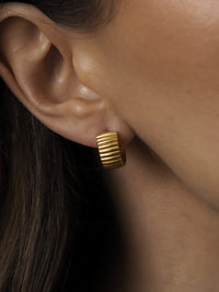 Close-up of a ridged gold hoop earring worn on the ear, showing its chunky, textured design.