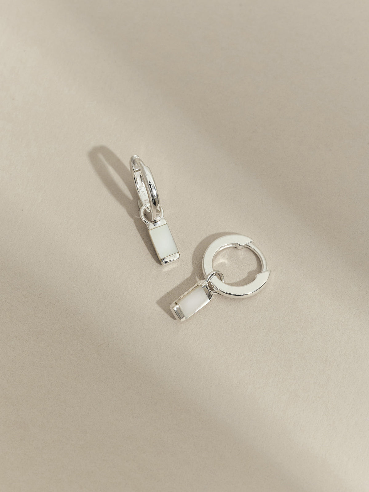 Flat lay of sterling silver hoop earrings with rectangular mother of pearl drop charms, displayed on a neutral beige surface.