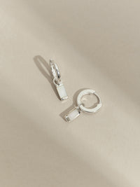 Flat lay of sterling silver hoop earrings with rectangular mother of pearl drop charms, displayed on a neutral beige surface.