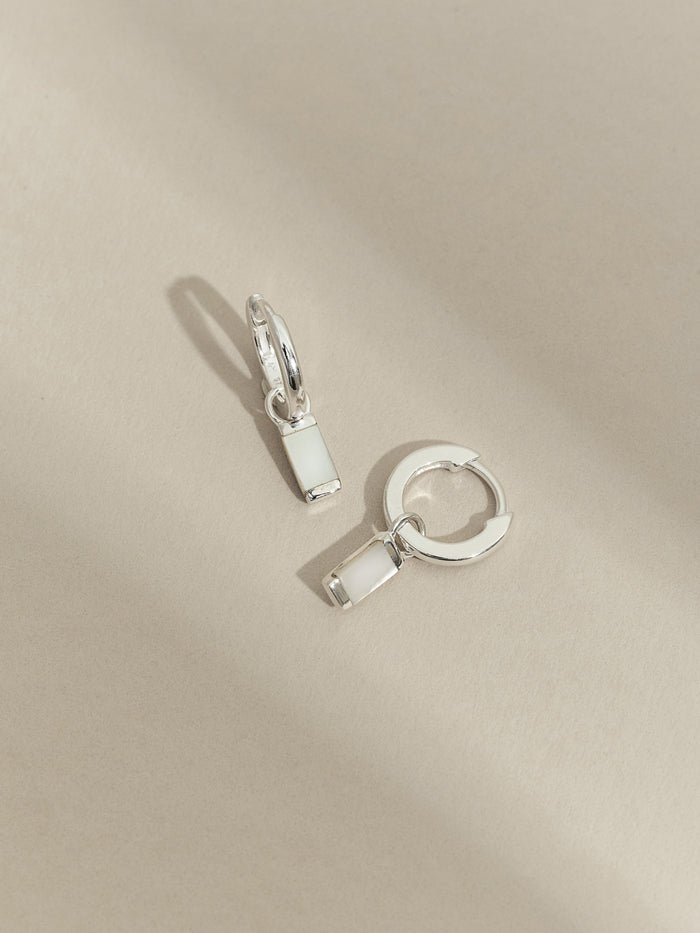 Flat lay of sterling silver hoop earrings with rectangular mother of pearl drop charms, displayed on a neutral beige surface.