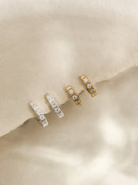 Two pairs of huggie earrings—one in sterling silver and one in gold—each set with cubic zirconia stones, styled on textured cream paper.