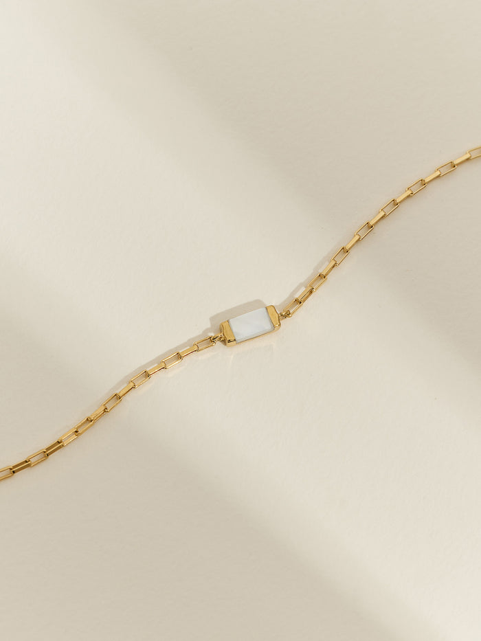 Close-up of a gold bracelet featuring a rectangular mother of pearl charm on a box chain, laid flat on a soft neutral background.
