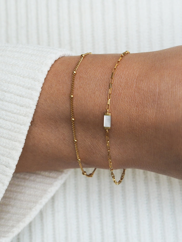 Gold bracelet gift set featuring a satellite chain and rectangular mother of pearl charm on a box chain, styled on a wrist against a white ribbed sleeve.
