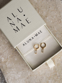 Gold hoop earrings with rectangular mother of pearl charms displayed in an Aluna Mae branded cream jewellery box, placed on a marble surface.