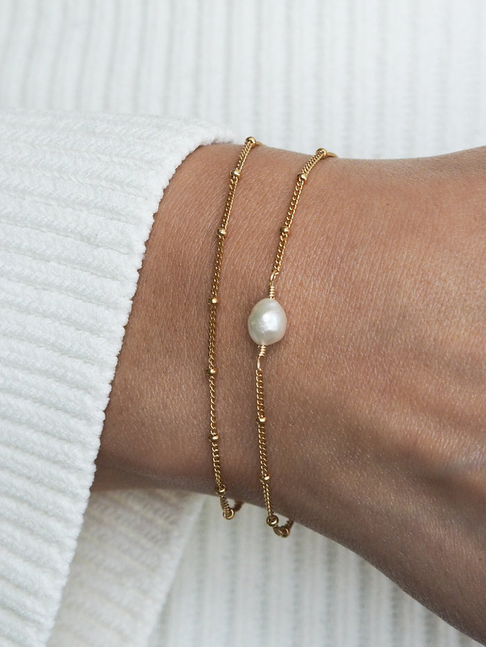 Gold satellite chain bracelet gift set featuring two 18ct gold plated sterling silver bracelets—one with a single freshwater pearl and one plain—styled on wrist with a white ribbed sleeve.
