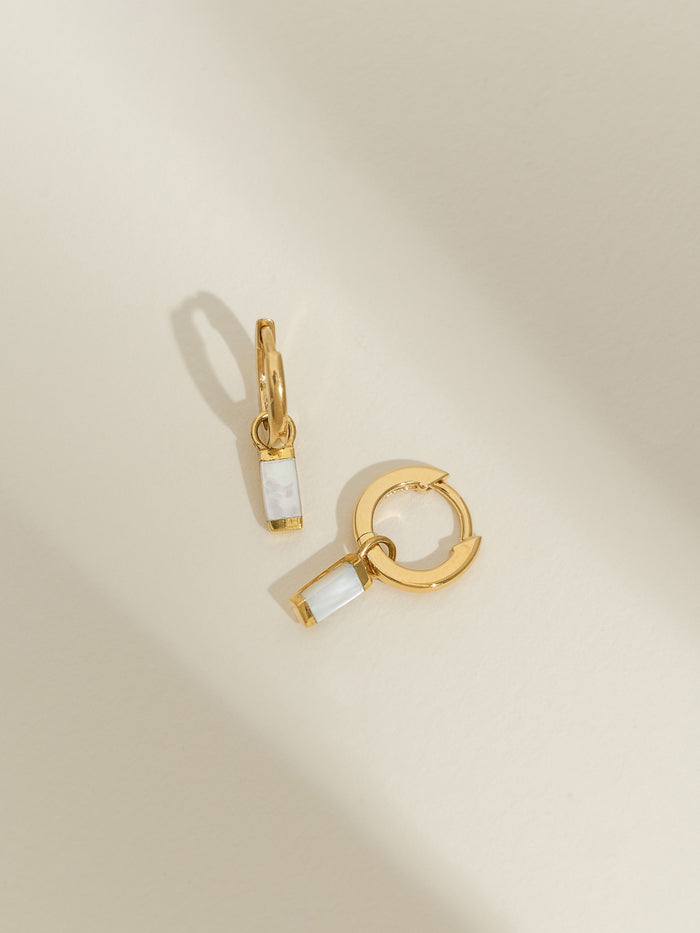 Gold-plated huggie hoop earrings with rectangular mother of pearl drop charms, displayed on a light cream background.