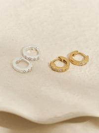 Two pairs of huggie earrings—one gold and one silver—each set with cubic zirconia stones, styled on a curved pale stone surface.