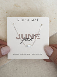 Hands holding a June birthstone bracelet featuring a rectangular mother of pearl stone on a sterling silver chain, displayed on a branded Aluna Mae card with the words 'Purity + Wisdom + Tranquility.
