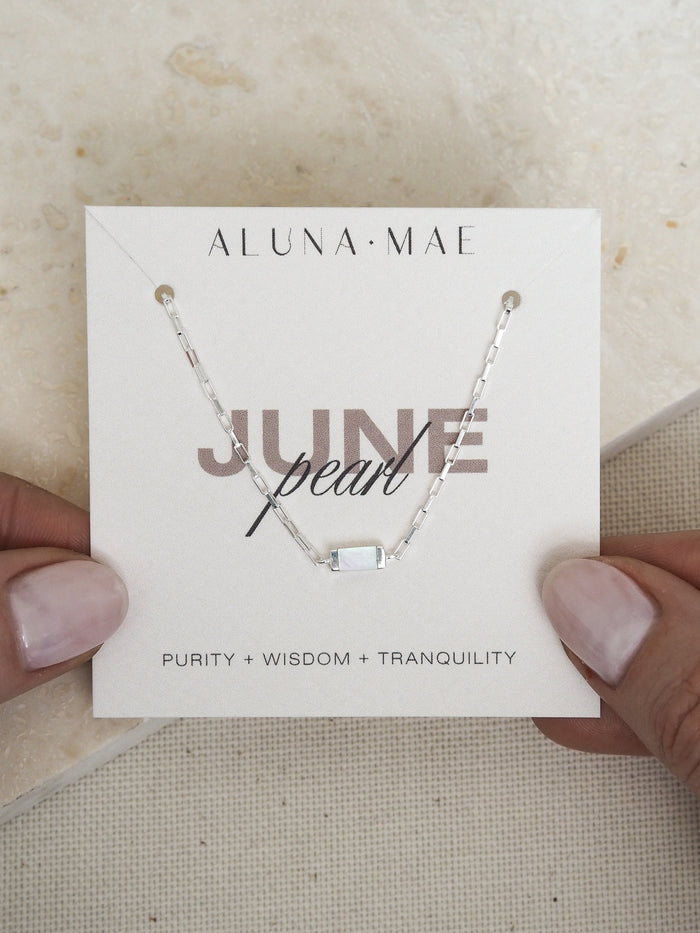 Hands holding a June birthstone bracelet featuring a rectangular mother of pearl stone on a sterling silver chain, displayed on a branded Aluna Mae card with the words 'Purity + Wisdom + Tranquility.