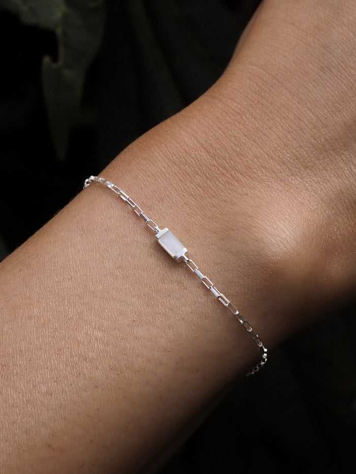 A close-up of a sterling silver box chain bracelet featuring a rectangular mother of pearl pendant, worn on a wrist with a dark blurred background.