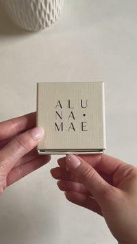 A hand opens an Aluna Mae jewellery gift box to reveal a bridesmaid message card inside.