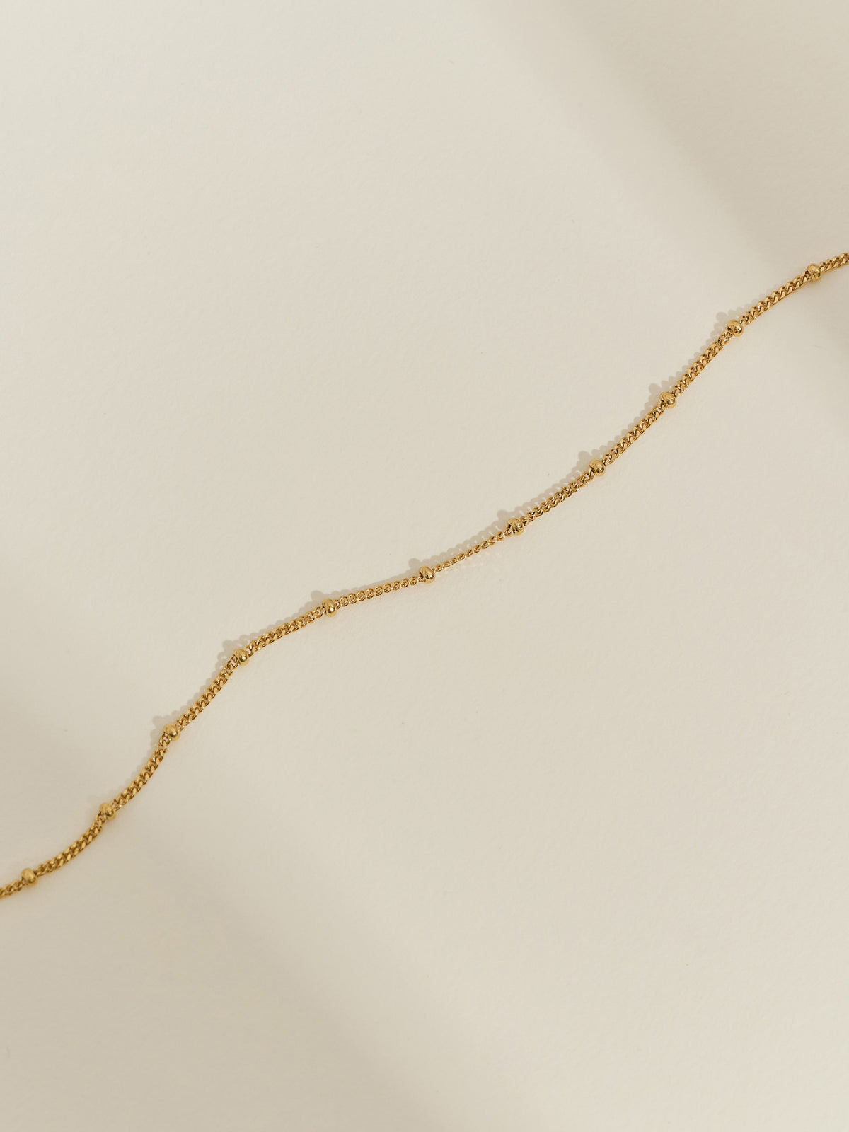 Minimal close-up of a gold-plated satellite chain necklace laid on a neutral background, showing its delicate beaded detailing.