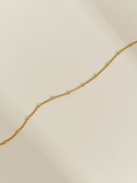 Minimal close-up of a gold-plated satellite chain necklace laid on a neutral background, showing its delicate beaded detailing.