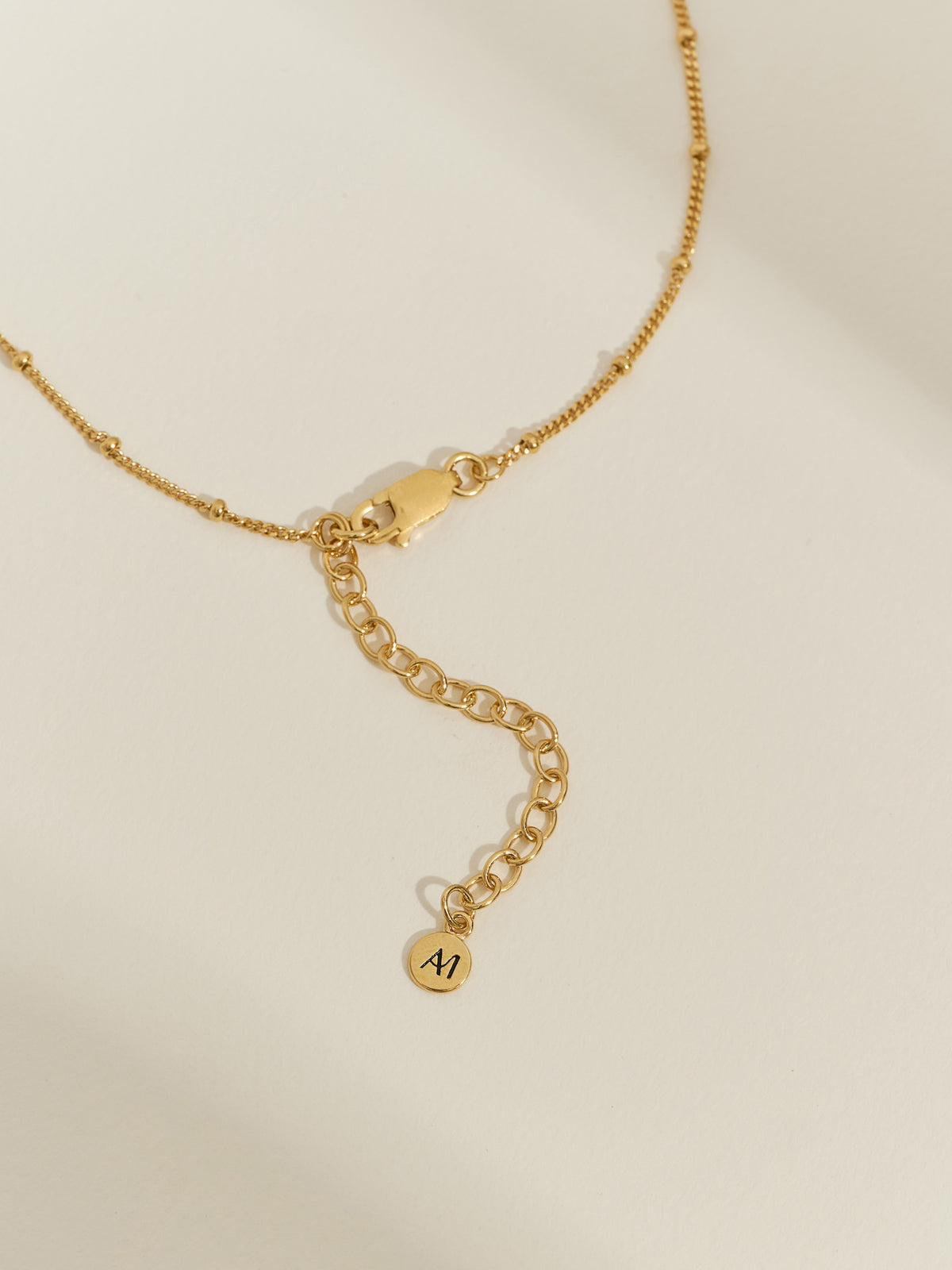 Close-up of a gold-plated necklace extender chain with lobster clasp and a small round Aluna Mae logo tag on a light background.