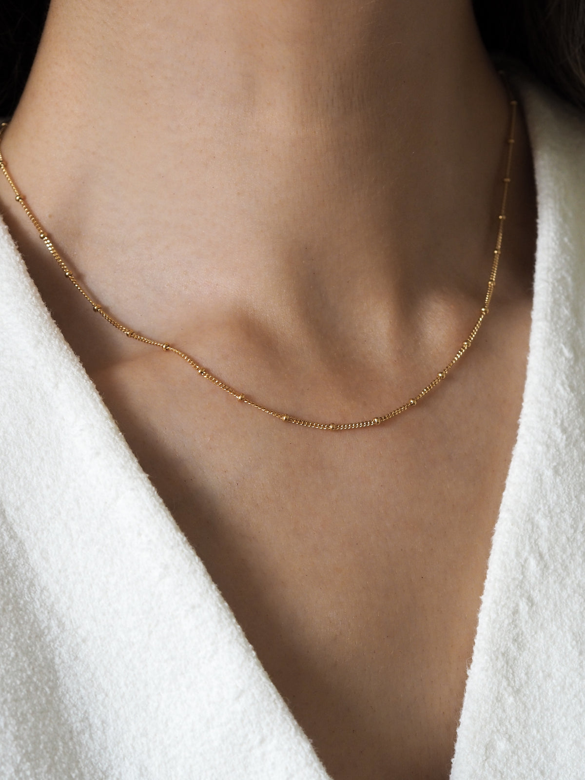 Close-up of a woman wearing a delicate gold-plated satellite chain necklace against a white textured top.