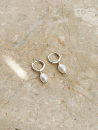A pair of sterling silver hoop earrings with oval freshwater pearl drops, displayed on a neutral-toned marble surface.