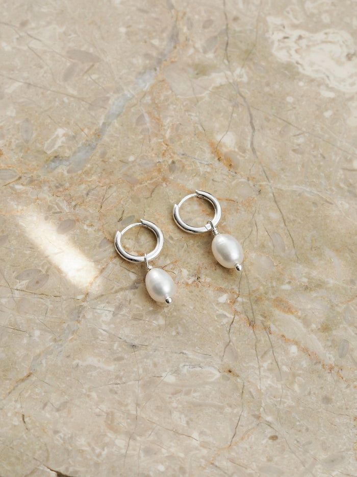 A pair of sterling silver hoop earrings with oval freshwater pearl drops, displayed on a neutral-toned marble surface.