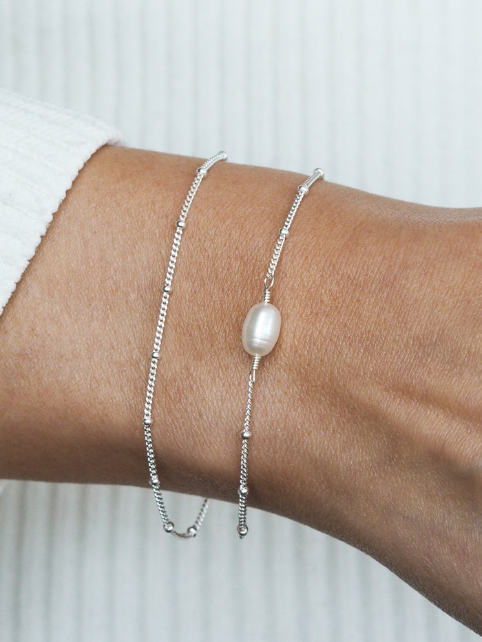 Close-up of a silver satellite chain bracelet gift set, featuring a single freshwater pearl on one bracelet, styled on a wrist with a ribbed white top.