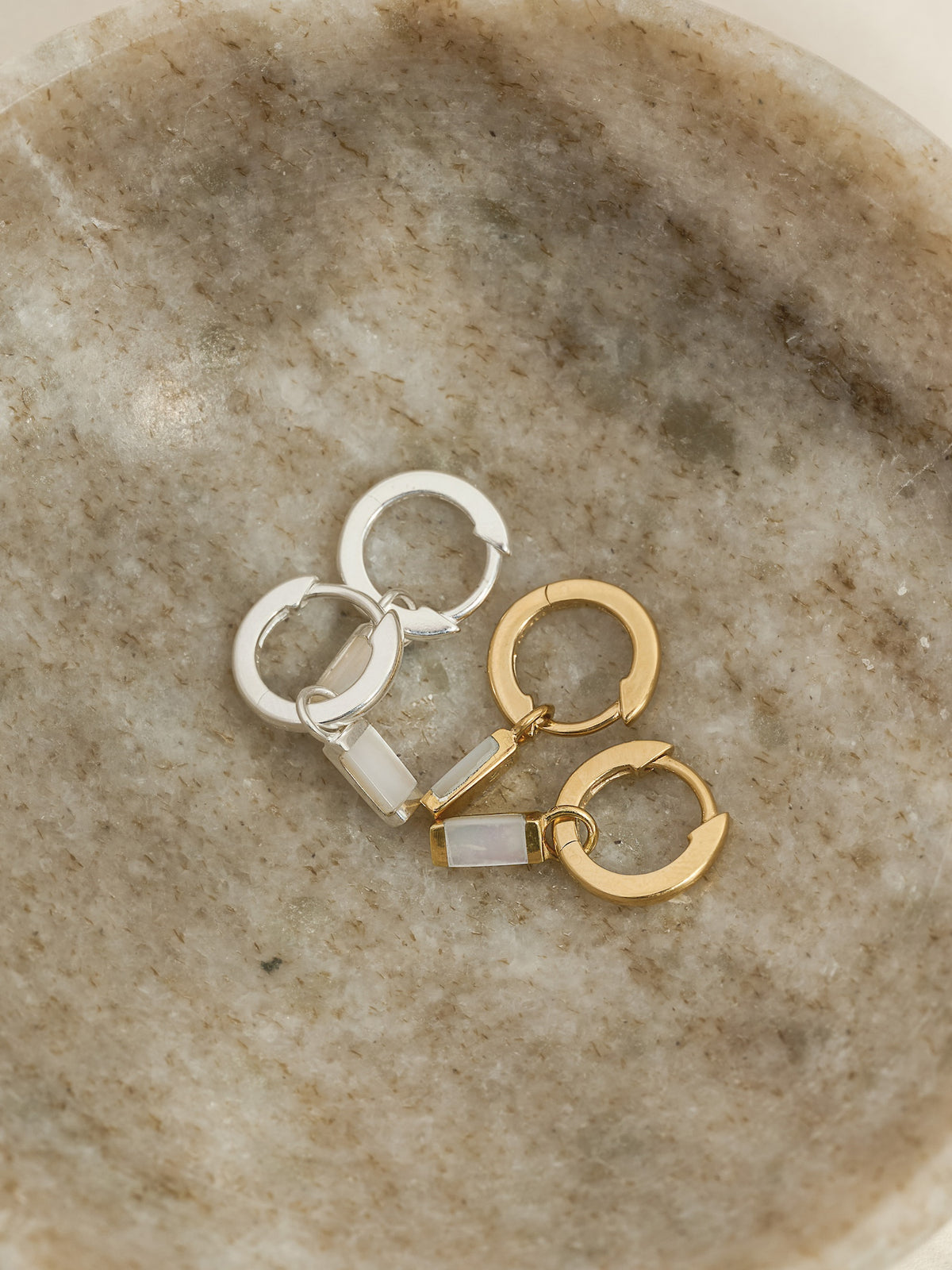 Close-up of two pairs of hoop earrings—one in sterling silver and one in gold—each featuring a rectangular mother of pearl drop charm, displayed in a shallow marble stone dish.
