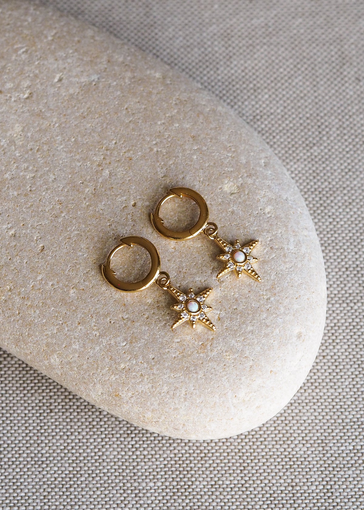 A pair of gold-plated huggie earrings with star-shaped opal and cubic zirconia charms, displayed on a smooth neutral-toned stone surface.