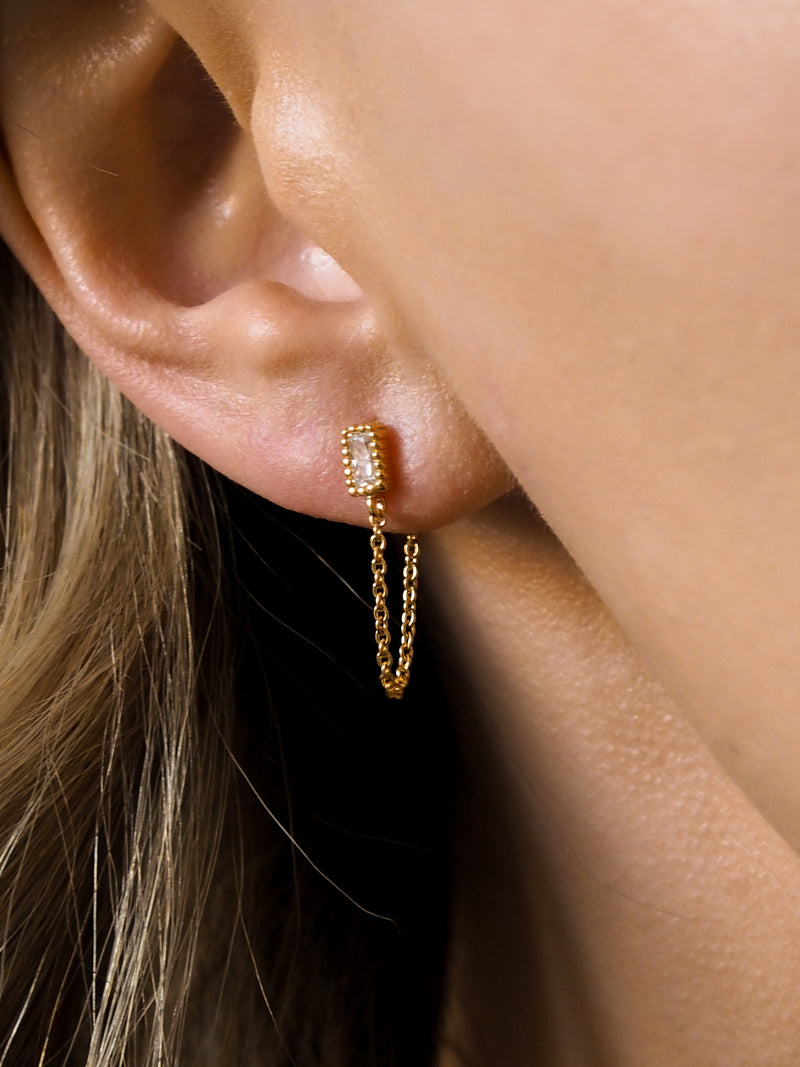 A model wearing gold stud earrings with chain and cubic zirconia gemstones.