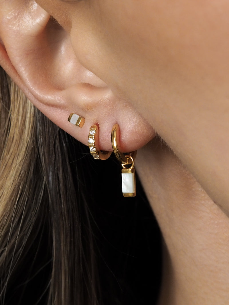 A model wearing multiple gold earrings layered together, featuring pearl pendant huggies, cubic zirconia huggies and mother of pearl studs. 