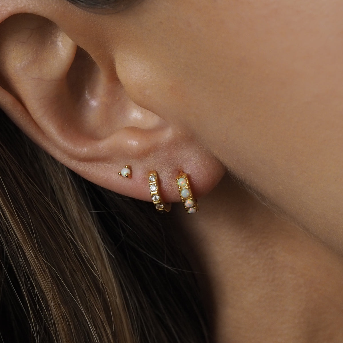 Close-up of an ear wearing a gold-plated opal stud and two gold-plated opal and cubic zirconia huggie earrings, styled for an ear stacking look.