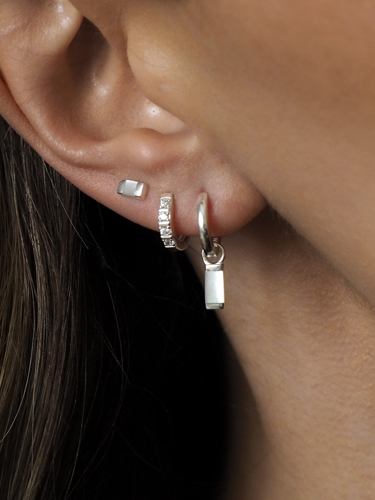 A model wearing multiple sterling silver earrings layered together, featuring mother-of-pearl pendant hoops, cubic zirconia huggies and mother-of-pearl studs. 