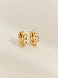 A pair of gold huggie earrings set with round opal stones, photographed against a soft cream background.