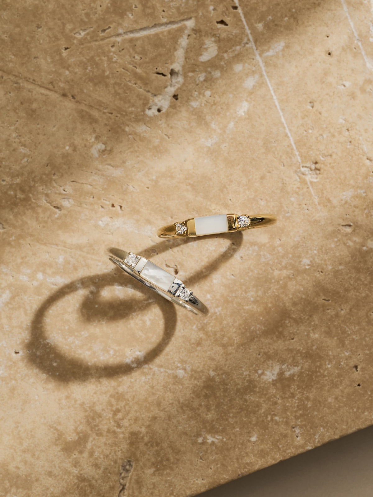 Two Aluna Mae rings displayed on travertine stone—one gold and one silver, each featuring a rectangular mother of pearl centre set between two cubic zirconia stones.