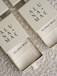 Multiple Aluna Mae jewellery gift boxes with the brand logo visible on the packaging. The boxes are open, revealing a fabric pouch inside with the brand name printed on it.