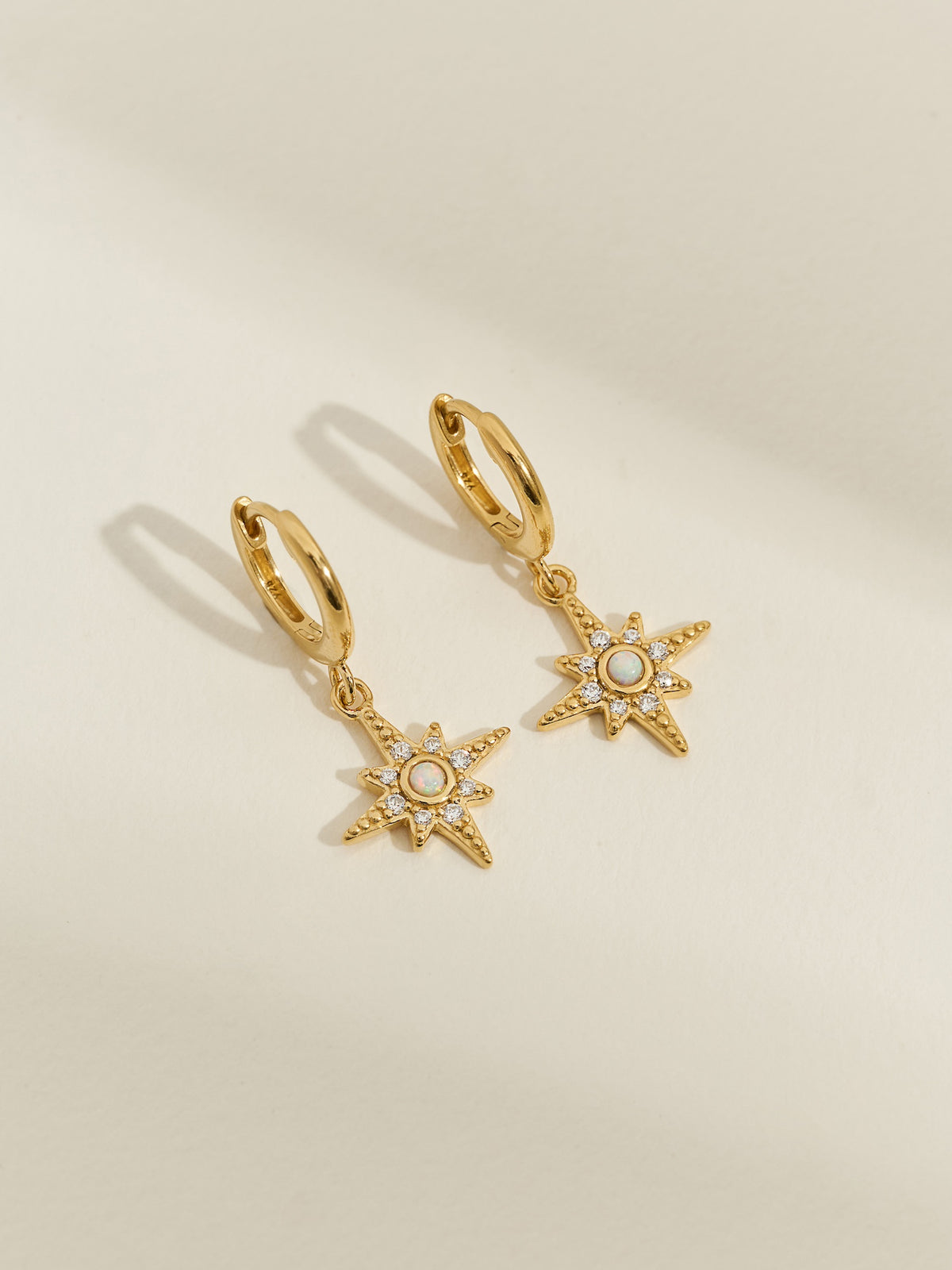 Close-up of gold star pendant hoops with opal and cubic zirconia gemstones against a plain backdrop.