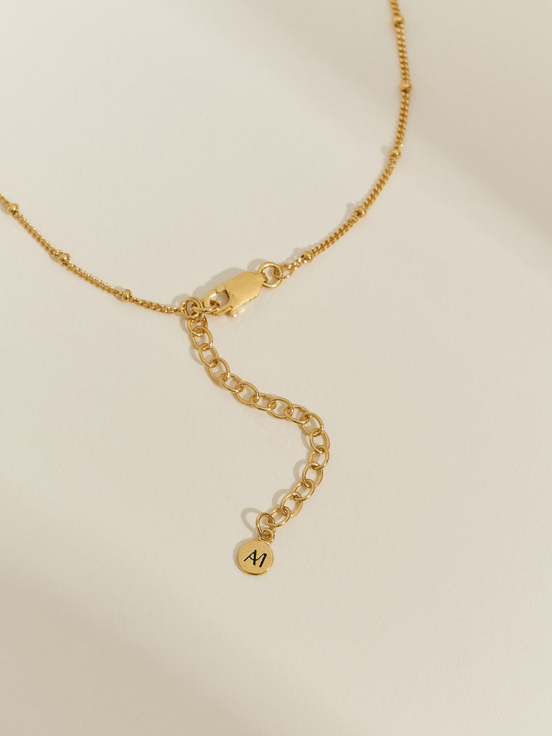 Close-up of a gold satellite chain bracelet with extender chain and Aluna Mae logo tag.