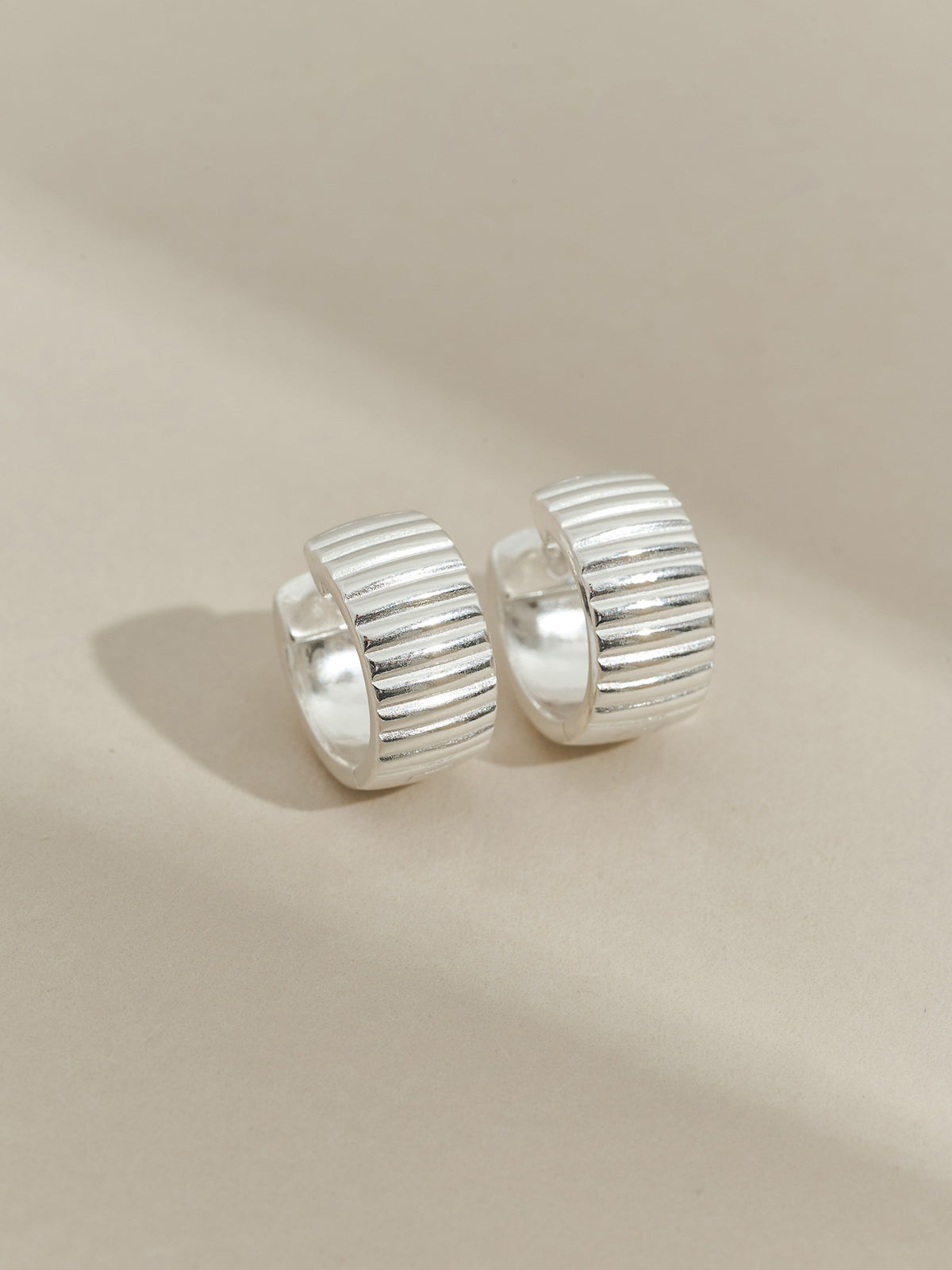 Pair of sterling silver ribbed hoop earrings displayed on a neutral beige background.