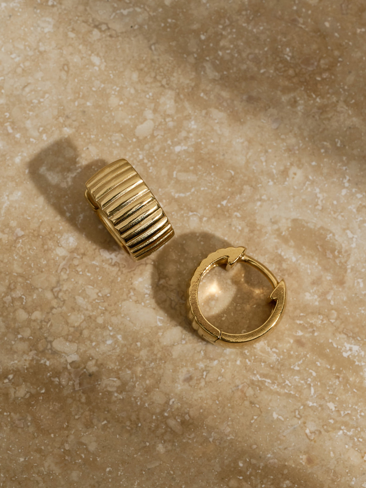 Pair of chunky ridged gold hoop earrings resting on a neutral-toned marble surface.