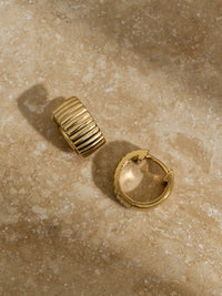 Pair of chunky ridged gold hoop earrings resting on a neutral-toned marble surface.
