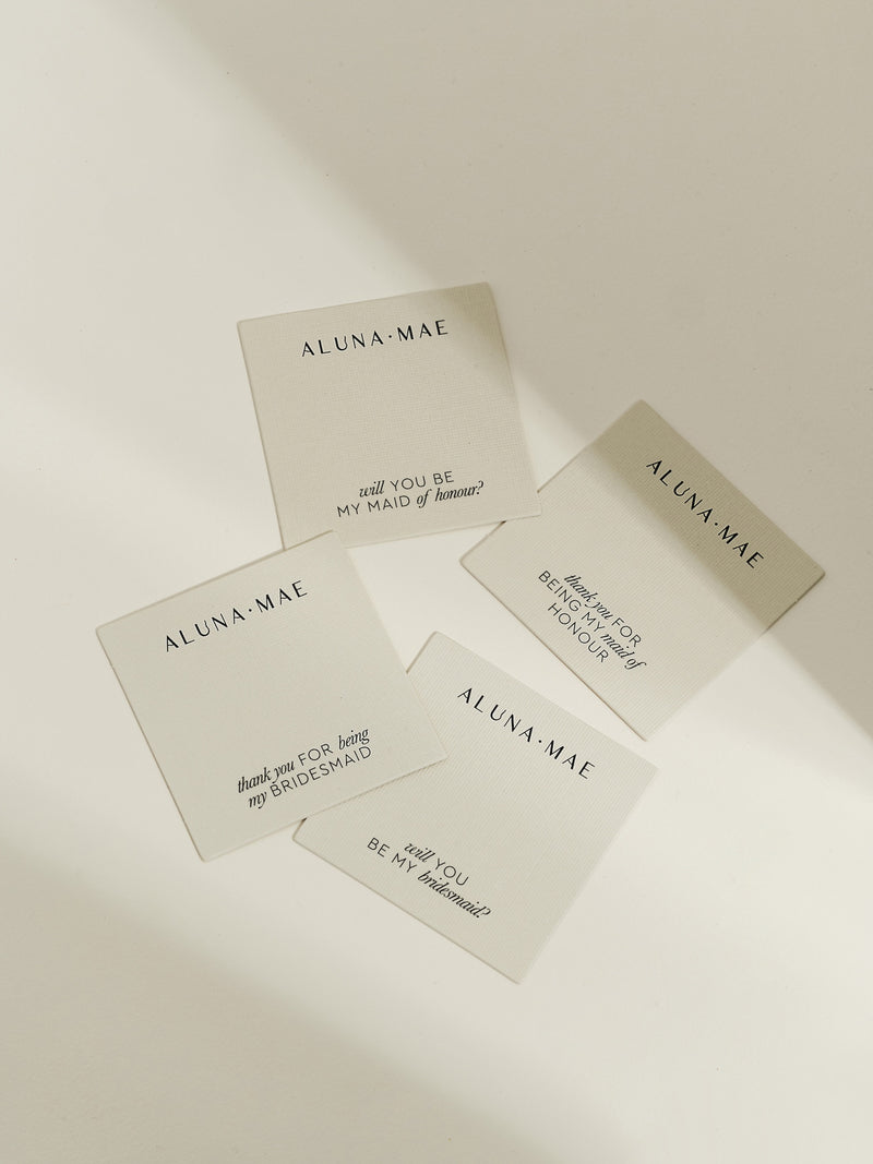 A flat lay of four Aluna Mae jewellery cards with wedding messages, including "Will you be my bridesmaid?" and "Thank you for being my maid of honour", displayed on a cream background in soft sunlight.