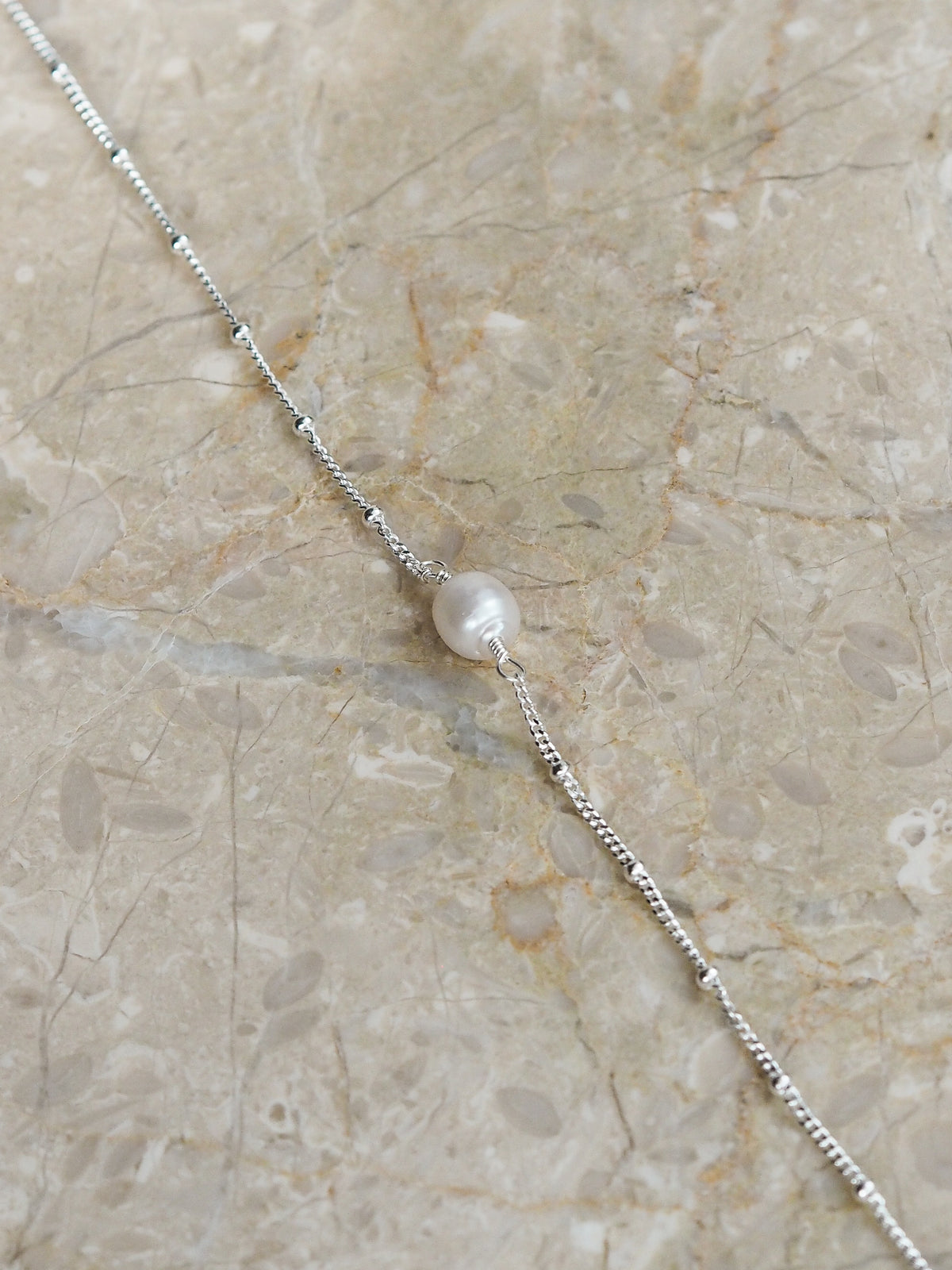 Flat lay of a sterling silver satellite chain bracelet featuring a single freshwater pearl, styled on a neutral marble surface.