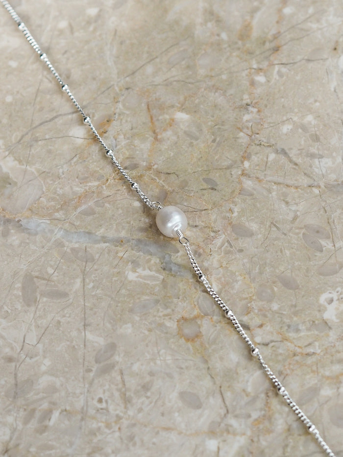 Flat lay of a sterling silver satellite chain bracelet featuring a single freshwater pearl, styled on a neutral marble surface.