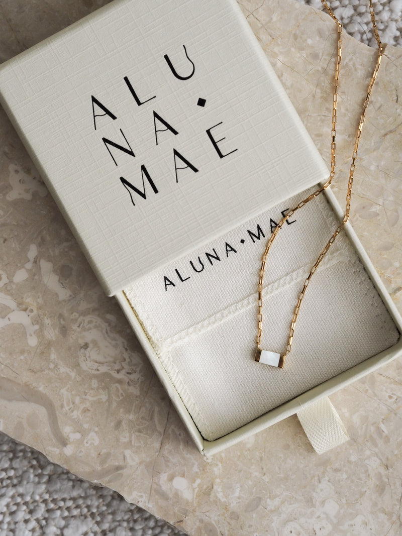 Gold box chain necklace with a rectangular mother of pearl pendant displayed in an Aluna Mae gift box on a travertine stone surface.