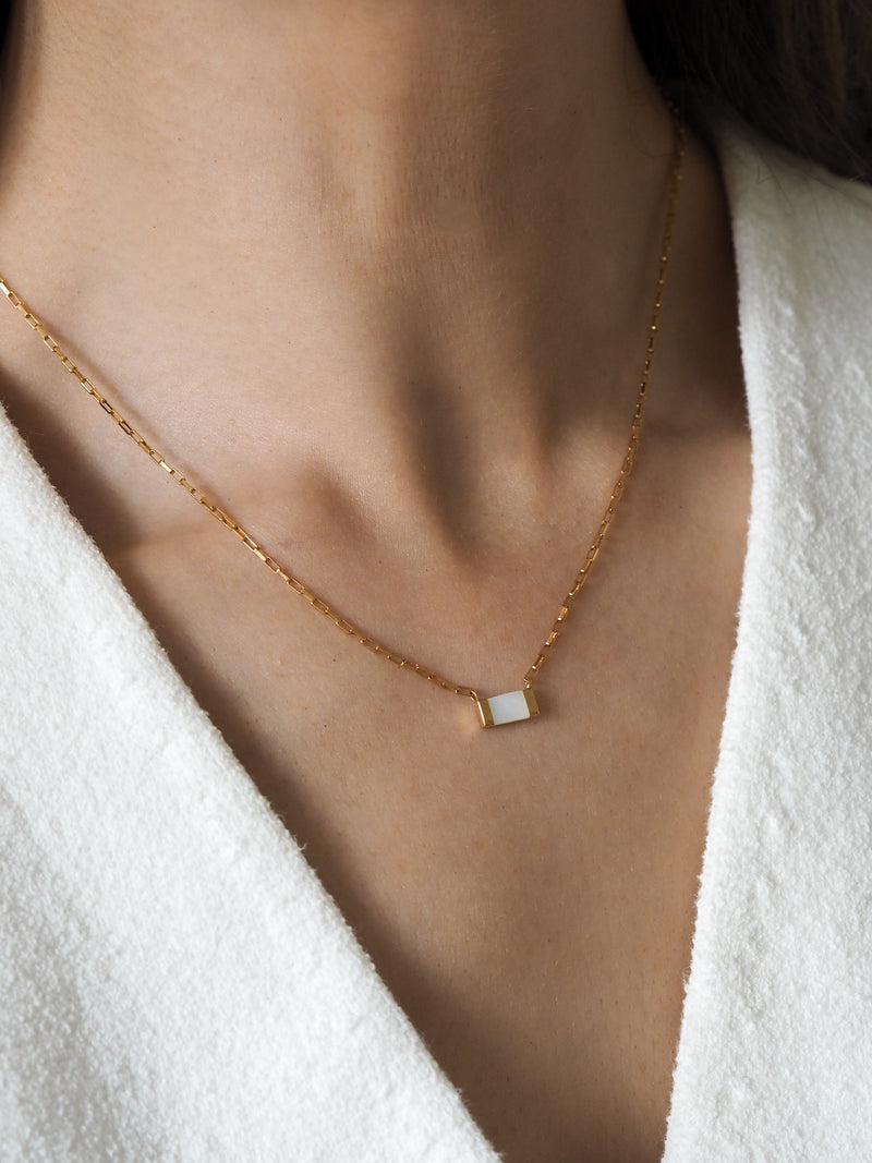 Gold box chain necklace with a rectangular mother of pearl pendant worn on model with a white textured fabric top.