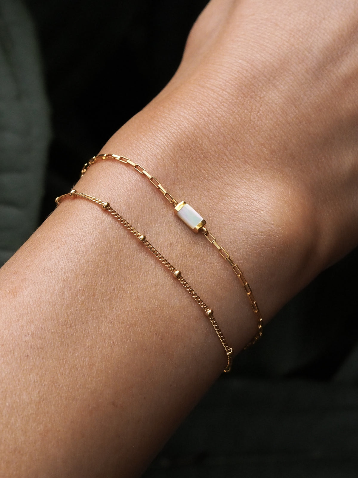 Two gold bracelets stacked on wrist—one features a rectangular mother of pearl stone on a box chain, the other is a delicate gold satellite chain bracelet.