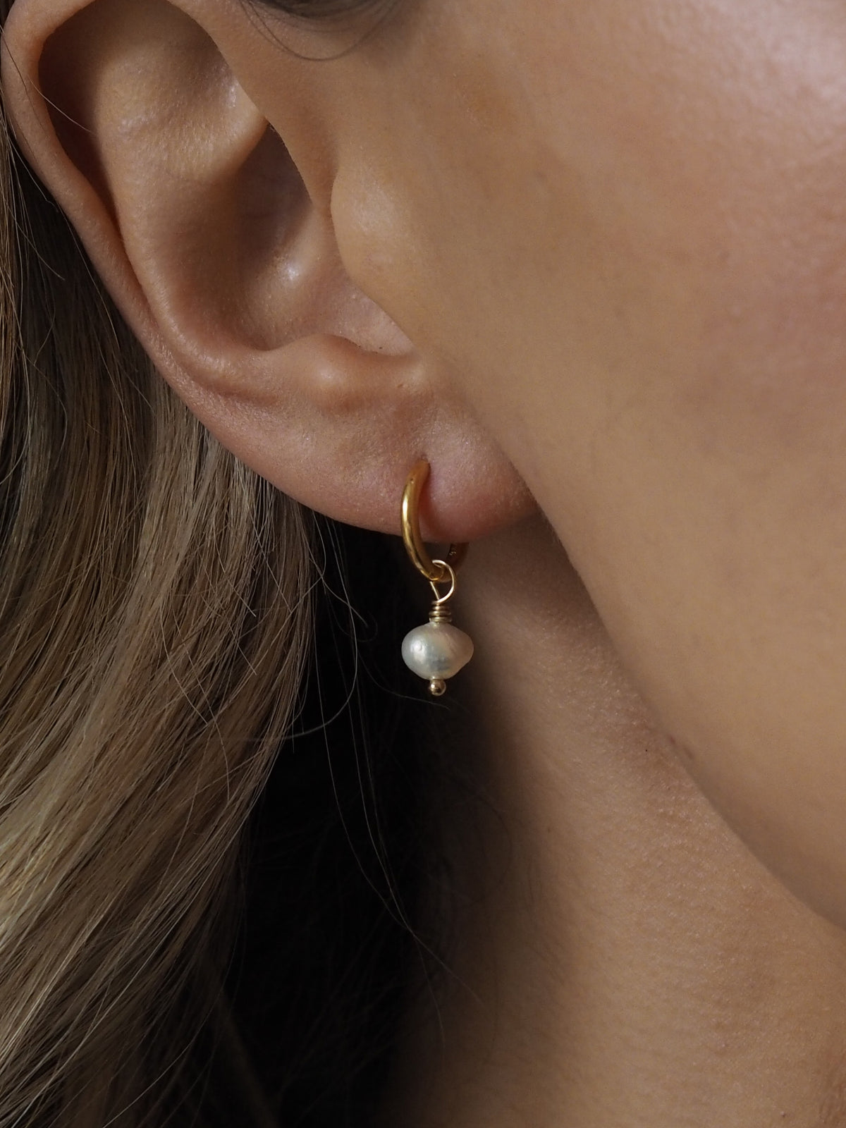 Close-up of a gold hoop earring with a single freshwater pearl drop, worn on an ear with blonde hair partially visible.