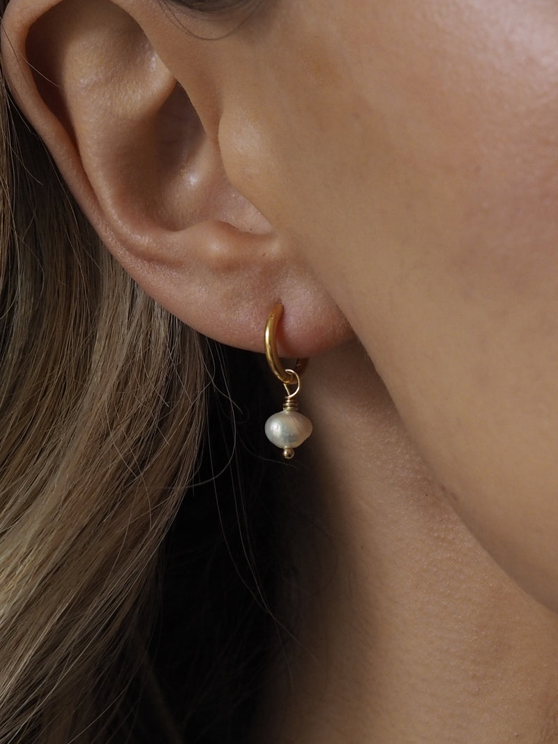 Close-up of a gold hoop earring with a single freshwater pearl drop, worn on an ear with blonde hair partially visible.